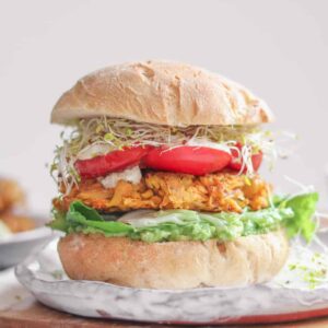 A vegan chickpea fritter burger with fresh vegetables and an avocado sauce sitting on a place before a white background