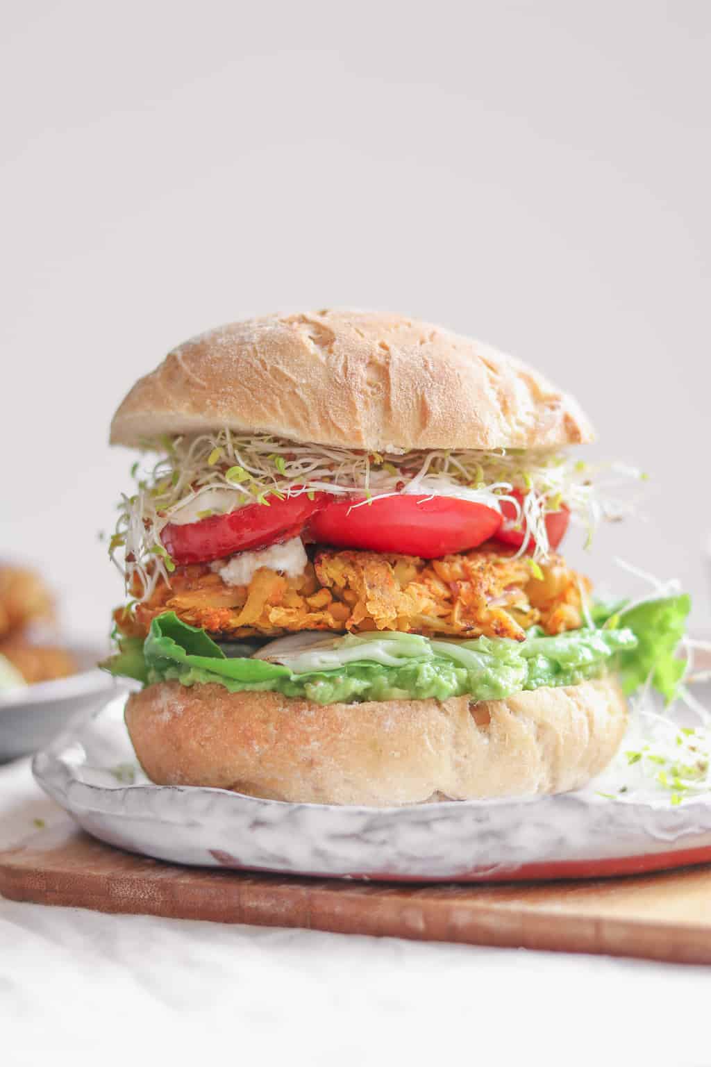 A vegan chickpea fritter burger with fresh vegetables and an avocado sauce sitting on a place before a white background